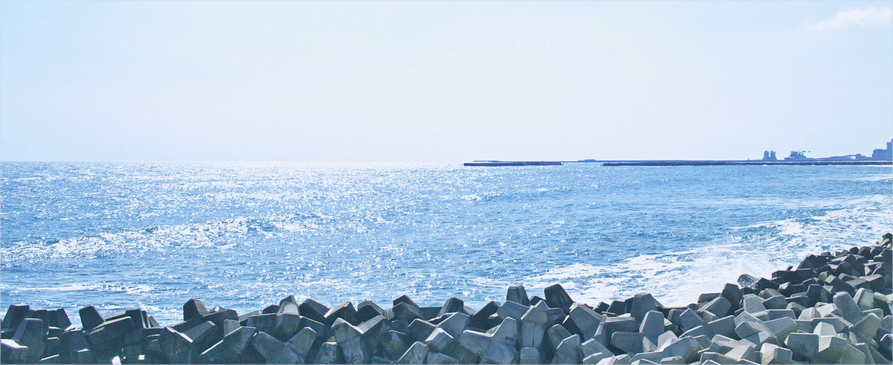 防波堤とテトラポッド越しに陽光を受けて輝く海が広がる風景。