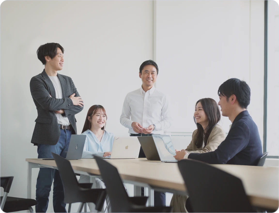 明るいオフィスで3人のビジネスパーソンがノートパソコンを囲んで打ち合わせをしている様子。中央の男性は白い長袖シャツを着て、ノートパソコンの画面を真剣に見つめ、右手を顎に当てて考えている。左に立つ女性はベージュのシャツを着て腕を組み、右方向を見ている。右に立つ男性は水色のシャツを着て、中央の男性の隣に立ち、笑顔で画面を見つめている。彼らは皆、集中して議論に参加しているように見える。