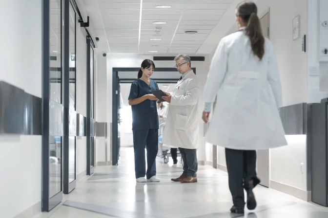 診察室で男性医師が女性患者に説明している様子。医師は白衣を着用し、右手を広げて説明しており、患者は穏やかな表情で耳を傾けている。二人の間には医療情報が表示された大きなモニターが置かれている。背景には大きな窓があり、自然光が差し込んでいる。