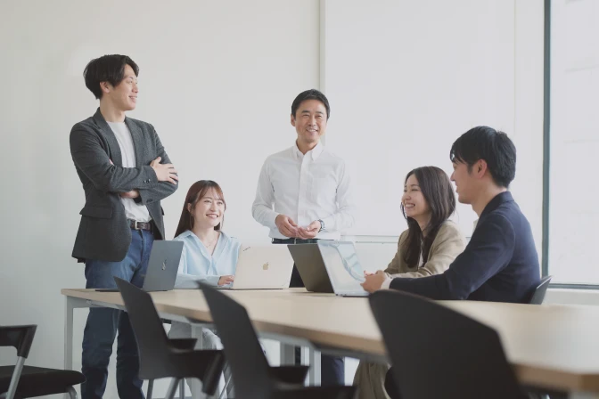 明るいオフィスで3人のビジネスパーソンがノートパソコンを囲んで打ち合わせをしている様子。中央の男性は白い長袖シャツを着て、ノートパソコンの画面を真剣に見つめ、右手を顎に当てて考えている。左に立つ女性はベージュのシャツを着て腕を組み、右方向を見ている。右に立つ男性は水色のシャツを着て、中央の男性の背後から画面を覗き込んでいる。開放的な窓のある空間で、活発な議論や共同作業が行われているような雰囲気。