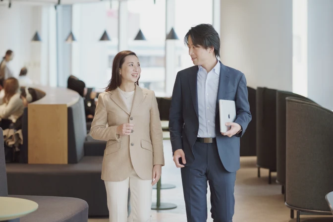 笑顔で会話しながらオフィスを歩く2人のビジネスパーソン。左側の女性はベージュのジャケットと白いインナーを着用し、右側の男性はネイビーのスーツと水色のシャツを着用し、左手にタブレットを持っている。2人は互いに顔を向け、楽しそうに話している様子。背景にはモダンなオフィス空間が広がり、窓から光が差し込んでいる。
