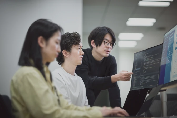 3人のアジア人開発者がオフィスで協力して作業している様子。中央の男性は白い長袖シャツを着て、左側からコンピューター画面を真剣に見つめている。右側の男性は黒い長袖シャツと丸い眼鏡をかけ、画面に表示されたコードを指差しながら説明している。画面にはダークモードでコードが表示されており、その手前にはノートパソコンが置かれている。左端には別の人物の肩と後頭部がぼやけて写っている。