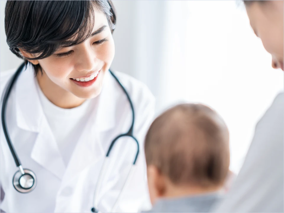 白衣を着用した笑顔の女性医療従事者が、背中を向けている赤ちゃんを見つめている。赤ちゃんの隣には、もう一人の大人の顔の一部が写っている。明るい光が差し込む診察室のような空間で、温かい雰囲気の医療現場の様子を捉えている。